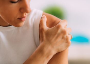 A person wearing a white tank top touches their right shoulder with their hand, looking thoughtful in a softly lit environment.