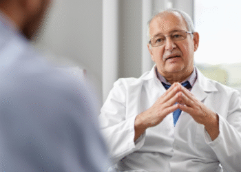 Un médico de mediana edad, con gafas y un bata blanca, está sentado frente a un paciente mientras habla y hace gestos con las manos en una consulta médica iluminada y moderna.