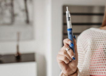 Una persona sostiene una pluma de insulina azul en un entorno interior, con una decoración minimalista en el fondo y una mano adornada con un anillo y uñas pintadas.