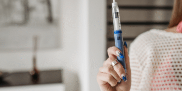 Una persona sostiene una pluma de insulina azul en un entorno interior, con una decoración minimalista en el fondo y una mano adornada con un anillo y uñas pintadas.