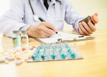 A doctor with a stethoscope dressed in a white coat holds a bottle of medication in one hand and writes on a form with the other, while vials and a variety of pills lie on the table.