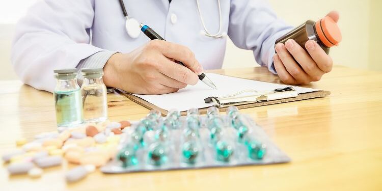 A doctor with a stethoscope dressed in a white coat holds a bottle of medication in one hand and writes on a form with the other, while vials and a variety of pills lie on the table.