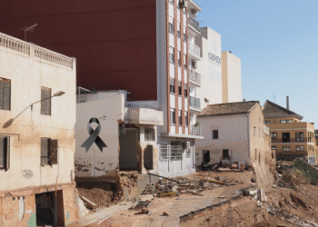Imagen de un área urbana dañada con edificios desmoronados y escombros visibles; se observa un lazo negro simbólico en el lado izquierdo.