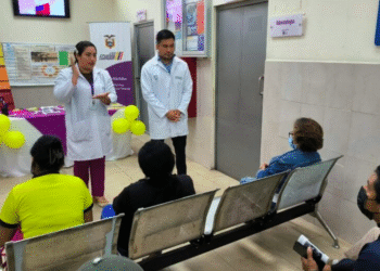 Deux professionnels de santé en blouse blanche s'adressent à un groupe de personnes assises dans une salle d'attente. Des ballons jaunes décorent la table en arrière-plan et des affiches informatives sont placardées sur les murs.