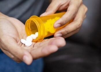 One hand holds an orange bottle of medicine, while another hand receives white pills that fall from the bottle into the palm.