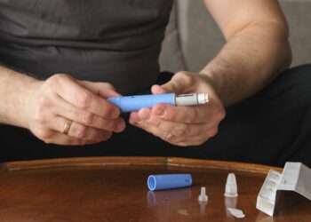 Una persona sostiene un dispositivo de inyección azul en sus manos, con un cartucho adicional y varias agujas desechables sobre una mesa de madera.