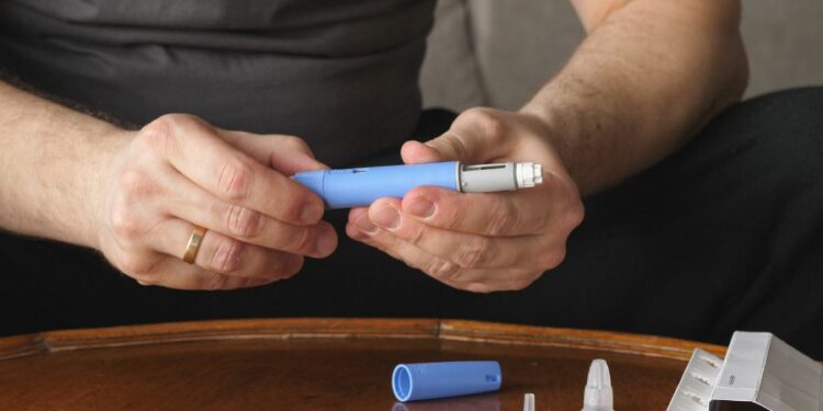 Une personne tient dans ses mains un dispositif d'injection bleu, avec une cartouche supplémentaire et plusieurs aiguilles jetables sur une table en bois.