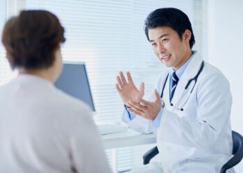 Un médico de cabello oscuro, vestido con una bata blanca y un estetoscopio alrededor de su cuello, está conversando animadamente con una paciente en un entorno moderno de consulta médica, con una computadora y ventanas iluminadas al fondo.