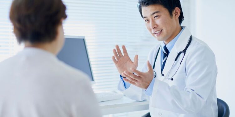 Un médico de cabello oscuro, vestido con una bata blanca y un estetoscopio alrededor de su cuello, está conversando animadamente con una paciente en un entorno moderno de consulta médica, con una computadora y ventanas iluminadas al fondo.