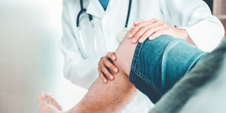 Un médico examina la rodilla de un paciente mientras ambos están sentados. El médico viste una bata blanca y tiene un estetoscopio alrededor del cuello.