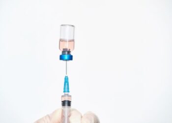 A gloved hand holding a syringe as it is filled with liquid from a clear vial containing a pink liquid.
