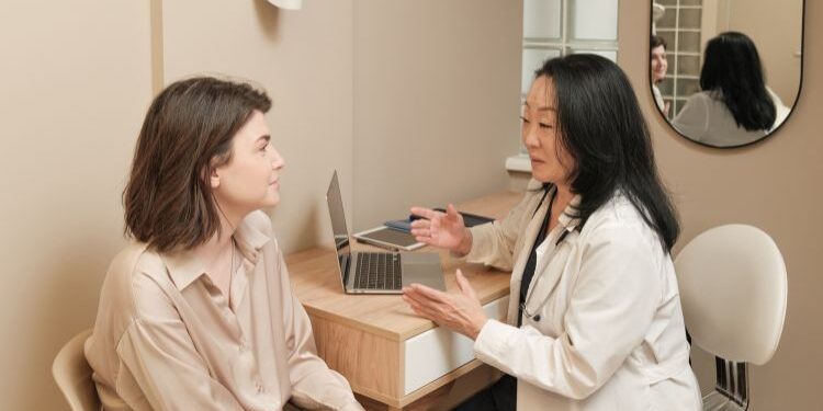 Una mujer con cabello castaño sentada, escuchando atentamente a una doctora de piel oscura con una bata blanca, que está hablando animadamente frente a una mesa con una laptop, en un consultorio moderno con paredes beige y un espejo circular en la pared.
