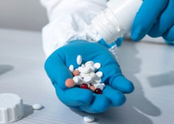 Una mano con guantes azules sostiene varias tabletas de diferentes colores, mientras otra mano vierte más pastillas de un frasco blanco en la palma.