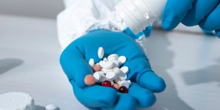 Una mano con guantes azules sostiene varias tabletas de diferentes colores, mientras otra mano vierte más pastillas de un frasco blanco en la palma.