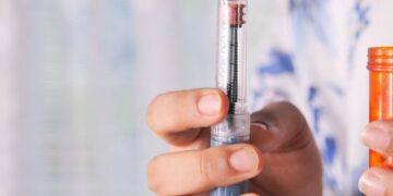 Una mano sostiene un dispositivo de inyección, mostrando una aguja y un indicador de dosis, mientras que en la otra mano se sostiene un recipiente anaranjado de medicación.