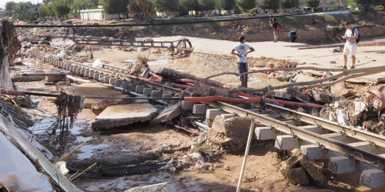 Imagen de un área afectada por inundaciones, con escombros, tubos y construcción dañada, y personas inspeccionando los restos.